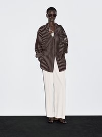 Braun-weißer gestreifter Oversize-Hemd, lockere weiße Hose, schwarze Sandalen und dunkle Sonnenbrille. Schlichte silberne Halskette, minimalistischer Handtasche.