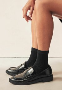 Musta nahkainen loafers-tyylinen kengät kiiltävällä pinnalla, joissa on pennin nauha, käytetty mustien ribbitempputossujen kanssa. Tasapohja ja näkyvä yksityiskohtainen ompelu.