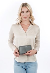 Woman with blonde hair wearing a beige blouse and light blue jeans holds a gray zippered wallet labeled "Tamaris" against a white background.