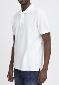 Vit polo-shirt med krage och två knappar, tillverkad av bomull. Har korta ärmar, sydd nederkant och en liten varumärkeslogotyp på bröstet.