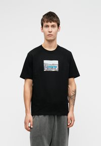 Tânăr cu părul scurt și închis la culoare, purtând un tricou negru cu un grafic de graffiti pe acoperiș și pantaloni gri, stând într-un fundal alb.