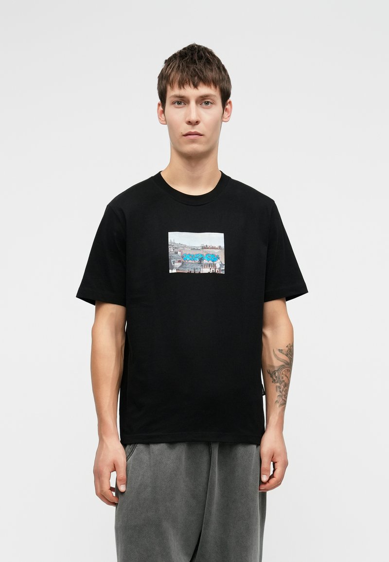 Tânăr cu părul scurt și închis la culoare, purtând un tricou negru cu un grafic de graffiti pe acoperiș și pantaloni gri, stând într-un fundal alb.