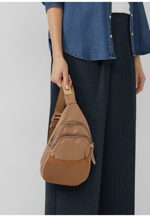 Braune Stoff-Schultertasche mit mehreren Reißverschlussfächern, Netzboden und einem hellbraunen Gurt. Wird auf der linken Schulter zu einer dunklen Hose getragen.