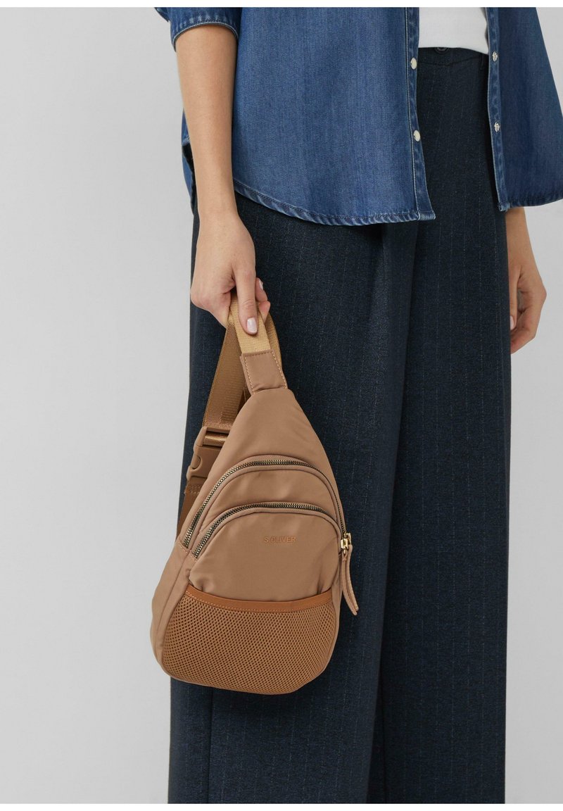 Borsa a tracolla in tessuto marrone con numerosi scomparti con zip, fondo in rete e tracolla beige. Indossata sulla spalla sinistra sopra pantaloni scuri.
