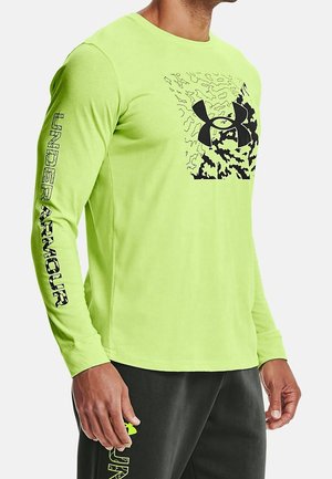 Homme portant un t-shirt vert citron à manches longues avec logo Under Armour noir, motif camouflage sur le devant et texte sur la manche gauche.