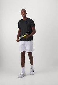 Mees, kes kannab musta Nike t-särki ja valgeid lühikesi pükse, hoides tennise reketit ja palli, seisab valgel taustal.