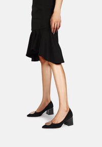 I décolleté in suede neri presentano una punta affusolata e un tacco a blocco, con un accento metallico sulla tomaia. L'outfit include una gonna nera con volant.