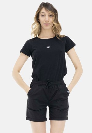 Femme aux cheveux blonds ondulés portant un t-shirt noir à manches courtes et un short noir, debout les mains dans les poches devant un fond blanc.