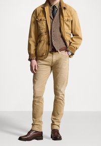 Tan bomulljacka med framfickor, brun herringbone-väst över en rutiga skjorta, beige chinos och bruna läderstövlar.