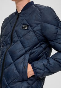 Marineblau steppierter Bomberjacke mit Reißverschluss, Seitentaschen, gerippten Bündchen und einem Logo-Patch auf der Brust. Glattes, strukturiertes Material.