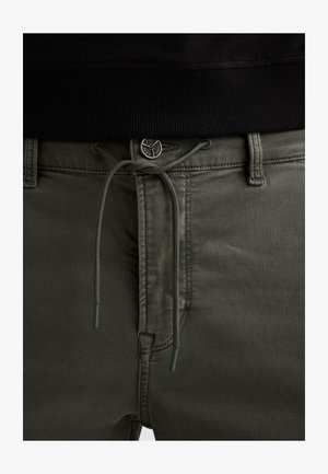 Krupni plan maslinasto zelenih hlača s vezicom vezanom u mašnu i metalnim gumbom s ugraviranim nazivom brenda iznad munje.