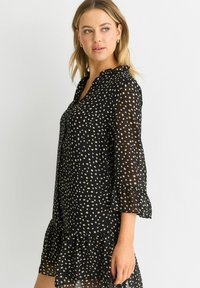 Black dress with a ruffled hem, featuring a sheer fabric and a pattern of beige dots. It has a V-neckline and three-quarter puff sleeves.