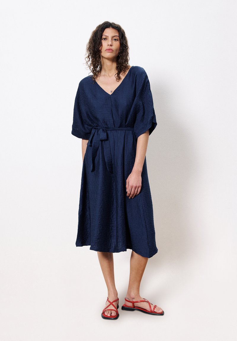 Robe bleu marine longueur genou en tissu texturé, avec un décolleté en V et des manches larges, ceinturée à la taille. Associée à des sandales rouges.