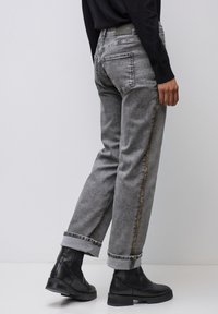 Jean en denim gris à coupe droite, avec une bande accent dorée le long du côté et des ourlets retroussés, porté avec des bottines noires.