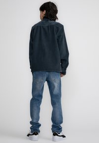 Tmavě modrá kordurová košile s kontrastním límcem, doplněná světle modrými džíny a černobílými teniskami. Pohled zezadu.