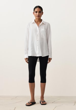 Woman standing wearing a white long-sleeve button-up shirt, black cropped leggings, and black flip-flop sandals on a light floor.