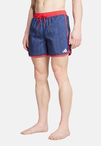 Niebieskie kąpielówki w wzory z czerwonymi akcentami, elastyczny pas i sznurek do regulacji. Zawierają logo Adidas po stronie dolnej. Lekki materiał.