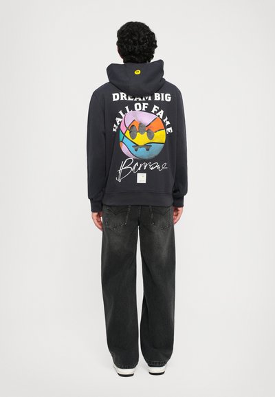Hoodie marine avec un design graphique coloré au dos, affichant le texte "RÊVE GRAND SALLE DE LA GLOIRE", associé à un jean ample de couleur foncée.