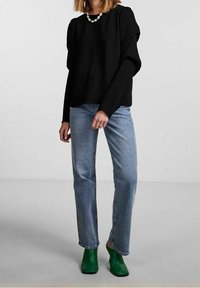 Schwarzes Langarmshirt mit herunterfallenden Schultern, kombiniert mit hellblauen Jeans und grünen Stiefeletten. Hat einen runden Halsausschnitt und eine strukturierte Silhouette.