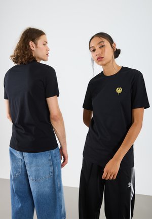 Camisetas de algodón negras en dos modelos, con un pequeño gráfico amarillo en el frente. Un modelo lleva pantalones cortos de mezclilla azules, mientras que el otro viste pantalones negros.