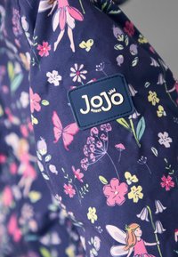 Laivyno spalvos audinys su gėlių ir fėjaus raštu pasteliniuose atspalviuose. Yra mėlynas etiketas su prekės ženklo pavadinimu "JoJo" baltomis raidėmis.