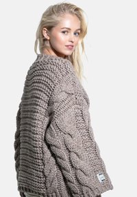 Beżowy dziergany sweter z grubym wzorem warkoczowym i prążkowaną fakturą, z szerokimi rękawami i naszywką przy dolnym brzegu.