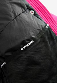 Čierna bunda vyrobená z lesklého nylonového materiálu, so sieťovanou kapsou a bielym logom "SUPERDRY". Na vrchu je viditeľná ružová rebrová časť.