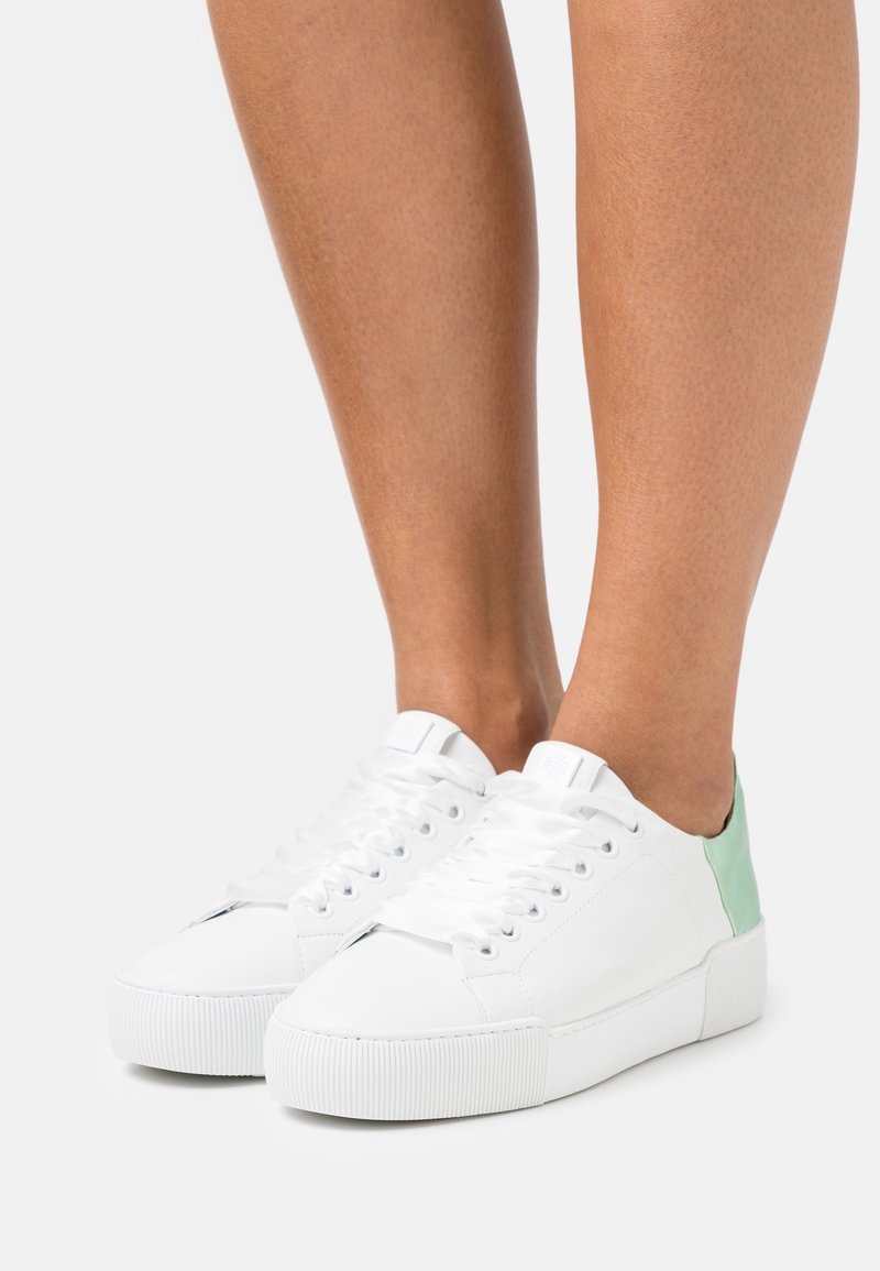 Jambes portant des baskets basses blanches avec des lacets blancs et une languette de talon vert clair sur un fond uni.