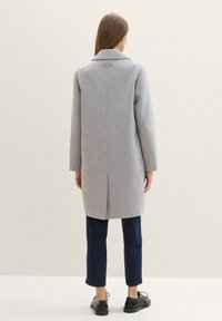 Manteau en laine gris avec un col en pointe et une fente arrière unique, porté sur un pantalon marine et des chaussures noires. Texture lisse et coupe ajustée.
