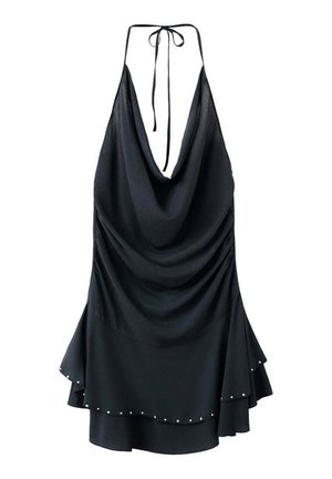 Jeune femme portant une robe noire sans manches avec un ourlet à volants et des sandales gladiateurs dorées, debout contre un mur blanc.