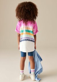 Przyduża biała koszulka z wielokolorowym wzorem tie-dye; różowe, niebieskie, pomarańczowe i zielone akcenty; zestawiona z niebieskimi szortami i białymi trampkami.