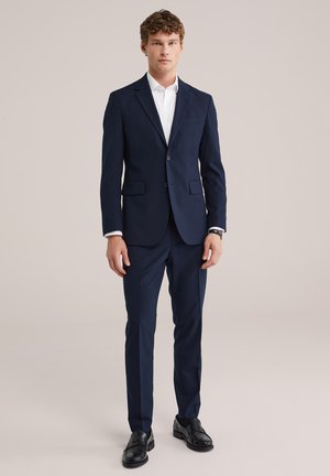 Giovane uomo che indossa un completo attillato blu navy con camicia bianca e scarpe eleganti nere, in piedi davanti a uno sfondo beige semplice.