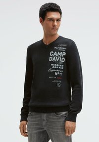Schwarzer gestrickter Pullover mit V-Ausschnitt, versehen mit weißem, gedrucktem Text und Akzenten. Gerippte Bündchen und Saum mit strukturierter Optik.