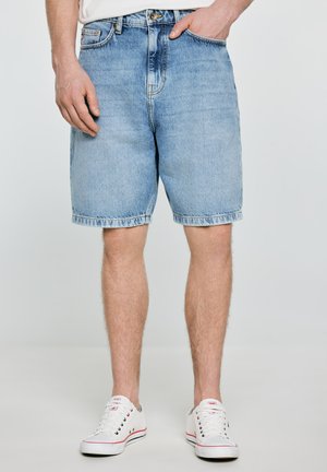 Mężczyzna w jasnoniebieskich jeansowych szortach i białych sneakersach, stojący z jedną ręką w kieszeni na jednolitym tle.