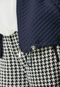 Dunkelblau gesteppte Jacke mit silbernem Reißverschluss und Kordelzug, kombiniert mit schwarz-weißen Hahnentritt-Musterhosen. Strukturierte und texturierte Stoffdetails.