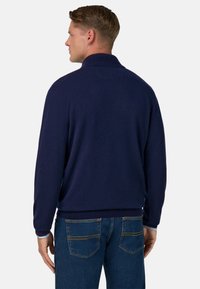 Dunkelblauer gestrickter Pullover mit Kragen, gerippten Bündchen und Saum, kombiniert mit blauen Jeans, die zwei Gesäßtaschen und gelbe Nähte haben.