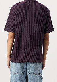 Fialové žebrované tričko s krátkým rukávem a texturovaným povrchem, s volným střihem a rovnou spodní hranou. Pohled zezadu ukazuje design límce.
