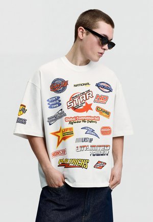 Mladi moški z očali za sonce in belo preveliko majico s pisanimi motivacijskimi in dirkaško obarvanimi tekstovnimi značkami, stoji z rokami v žepih.