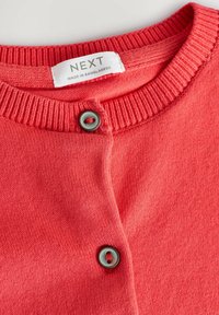 Cardigan en tricot corail avec un col côtelé, présentant une fermeture à boutons gris et une étiquette indiquant "NEXT, FABRIQUÉ AU BANGLADESH." Texture lisse.