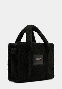 Sac noir avec un extérieur texturé et flou, doté de poignées courtes et d'un patch logo noir mat. Forme carrée avec des détails cousus.