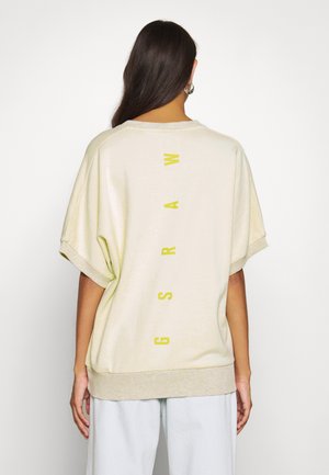 Vrouw met lang donker haar, gekleed in een losse crèmekleurige top met korte mouwen, met gele verticale "GSRAM" letters op de achterkant.