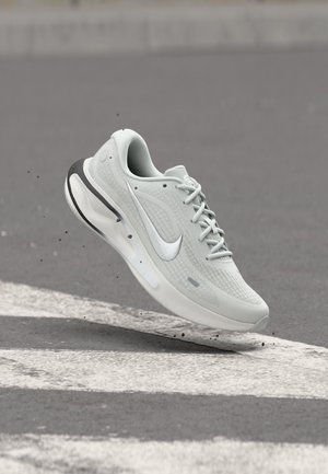 Witte Nike hardloopschoen met zichtbare luchtkussen, zwevend in de lucht boven asfalt met witte geschilderde lijnen en kleine stukjes vuil eromheen.