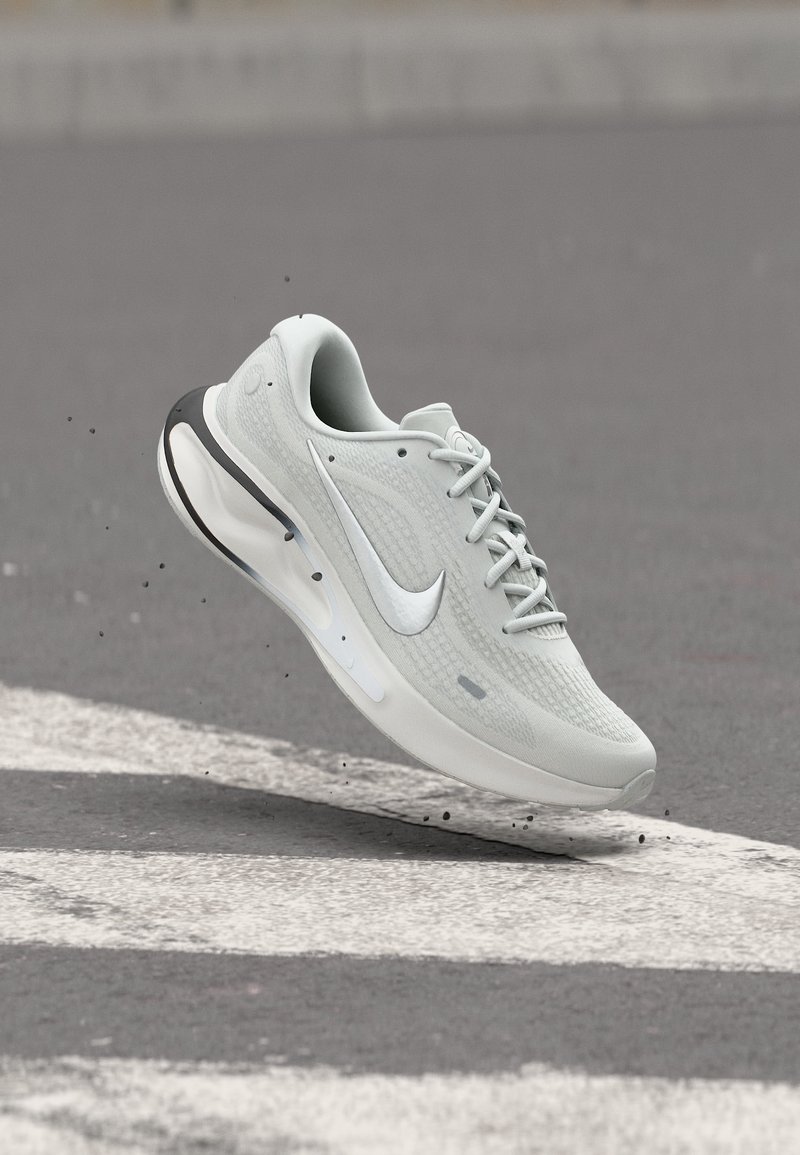 Λευκό αθλητικό παπούτσι Nike για τρέξιμο με ορατό αερόσακο, στον αέρα πάνω από άσφαλτο με λευκές γραμμές ζωγραφισμένες και μικρά υπολείμματα γύρω του.