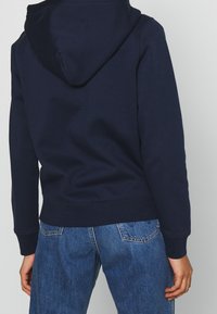 Felpa blu navy realizzata in tessuto morbido, dotata di cappuccio con cordino e vestibilità comoda. Abbinata a jeans blu, mostrata dalla parte posteriore con polsini a costine.