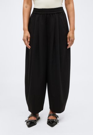 Henkilö pukeutuneena löysiin mustiin leveälahkeisiin housuihin, joissa on joustava vyötärönauha, ja mustiin niitattuihin teräväkärkisiin tasapohjaisiin kenkiin valkoisella taustalla.