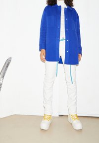 Blå ullkappa med framfickor, vit höghalsad skjorta, vita byxor med turkosa detaljer och vita sneakers med gula snören.