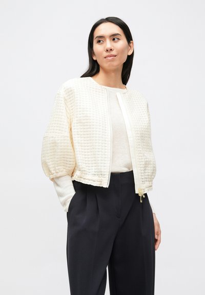 Κρεμ cropped μπουφάν με υφασμάτινο μοτίβο, εμπρόσθιο φερμουάρ κλεισίματος και ριμπ μανσέτες, συνδυασμένο με σκούρο ψηλόμεσο παντελόνι.