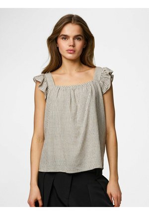 Młoda kobieta ubrana w beżową, bezrękawnikową bluzkę w kratę z falbankowymi ramiączkami i czarne spodnie, stojąca na jasnym, jednolitym tle.