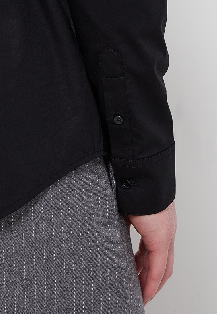 Chemise noire à manches longues avec des poignets boutonnés, présentant une texture lisse et une coupe structurée. Portée sur un pantalon gris à rayures.