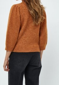 Mujer con cabello largo vistiendo un suéter de canalé en color naranja quemado con mangas abullonadas y jeans negros de cintura alta, mostrada de espaldas.